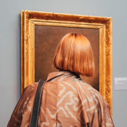 Person with blonde bob stands with back to camera, looking at a framed painting in a gallery room