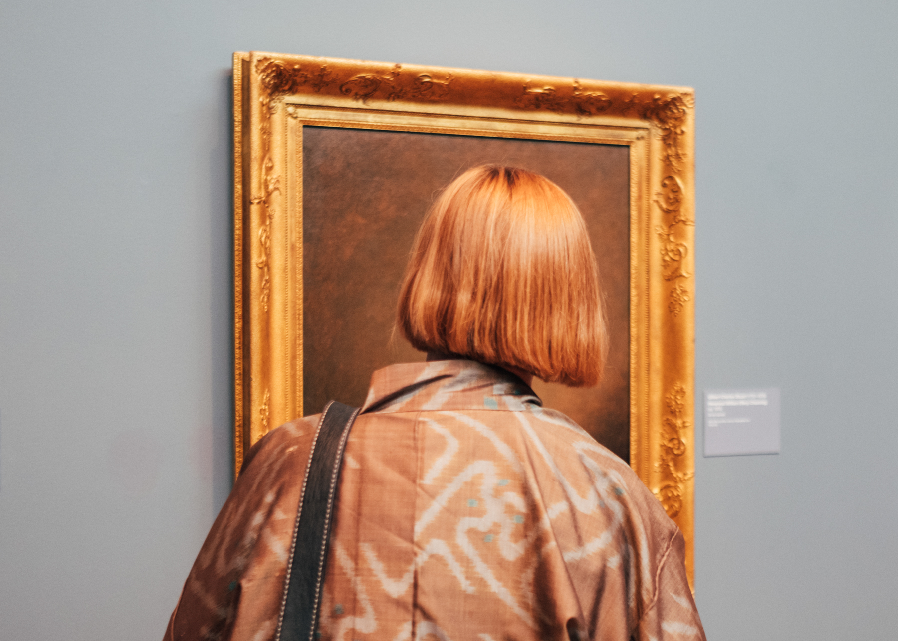 Person with blonde bob stands with back to camera, looking at a framed painting in a gallery room