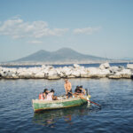 Group of people in a small green rowboat rowing on calm water near a rocky breakwater with distant mountains under a blue sky.