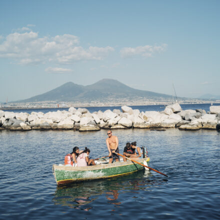 Workshop photo à Naples – Construire son regard Group of people in a small green rowboat rowing on calm water near a rocky breakwater with distant mountains under a blue sky.