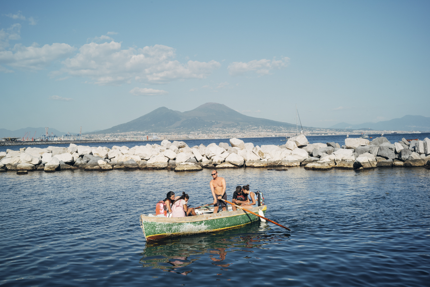 Group of people in a small green rowboat rowing on calm water near a rocky breakwater with distant mountains under a blue sky.